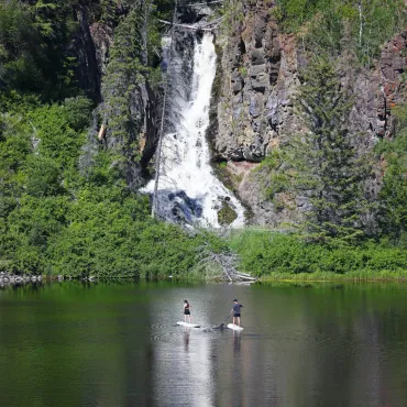A couple paddle board on a serene wilderness lake under a cascading waterfall